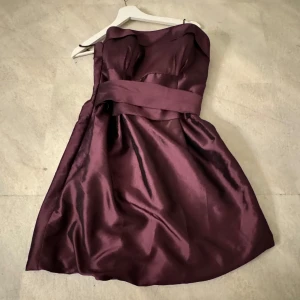 Lila axelbandslös klänning - Elegant klänning i glansigt material med axelbandslös design. Den har en markerad midja och veckad kjol som ger en fin siluett. Perfekt för festliga tillfällen.