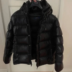 Svart dunjacka från Polo Ralph Lauren - Snygg svart dunjacka från Polo Ralph Lauren med dragkedja och knappar. Jackan har en pufferdesign och är perfekt för kyliga dagar. Nästan inte använd.
