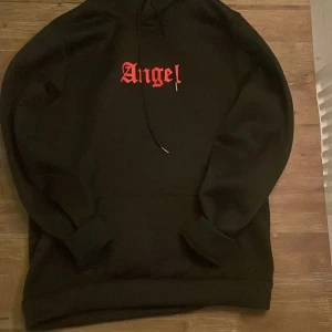 Svart hoodie med röd text - Snygg svart hoodie med texten 'Angel' i rött på både framsidan och baksidan. Perfekt för en avslappnad stil med en touch av attityd. Den har en klassisk passform med huva och dragsko.