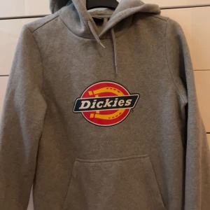 Grå hoodie från Dickies - Säljer en klassisk grå hoodie från Dickies med deras ikoniska logga på bröstet. Tröjan har en bekväm passform med känguruficka och justerbar huva. Perfekt för en avslappnad stil.