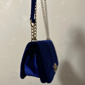 Blå quiltad axelväska i sammet - Snygg blå axelväska i sammet med quiltad design och guldfärgad kedja. Väskan har ett elegant spänne framtill och en praktisk storlek för att bära med sig det viktigaste. Perfekt för att ge en touch av lyx till din outfit.