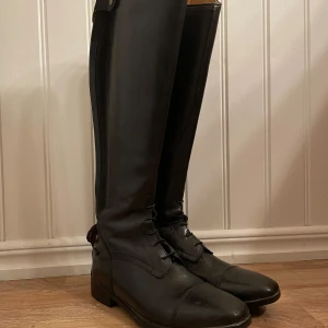 Svarta ridstövlar med dragkedja - Snygga svarta ridstövlar i skinn med dragkedja längs baksidan. De har en klassisk design med snörning framtill och en låg klack. Perfekta för ridning och ger ett stilrent intryck. Panaro CRW storlek 39. Passar perfekt för vad omkretsen 36-37 cm,  obetydligt använda. Säljes då de var för vida i vaden för dottern