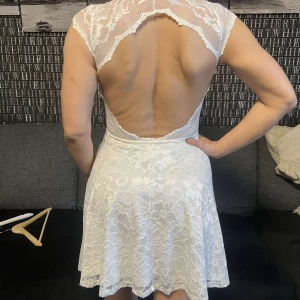 Vit spetsklänning med öppen rygg - Elegant vit spetsklänning med öppen rygg och korta ärmar. Klänningen har en vacker spetsdesign och en feminin siluett som passar perfekt för speciella tillfällen.