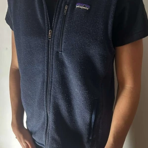 Mörkblå fleeceväst från Patagonia - Säljer en mörkblå fleeceväst från Patagonia med dragkedja och praktiska fickor. Perfekt för lager-på-lager under kyliga dagar. Västen har en stilren design med en liten logga på bröstet.