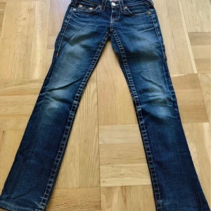 Blå jeans från True Religion - Snygga blå jeans från True Religion med klassisk femficksdesign och orange sömmar. Jeansen har en bootcut-stil och är tillverkade i USA. Perfekta för en avslappnad look.