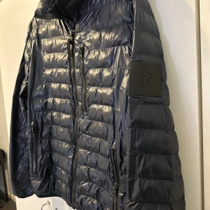 Svart pufferjacka från Peak Performance - Snygg svart pufferjacka från Peak Performance med dragkedja och flera praktiska fickor. Jackan har en quiltad design och är perfekt för kyligare dagar. Den har en normal passform och långa ärmar. på vänsta arm på baksidan fins det ett lite hål se bilden!