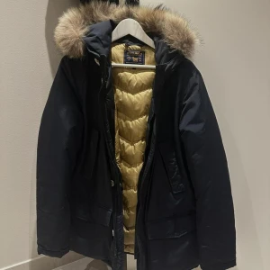 Svart parkajacka från Woolrich - Säljer en stilren svart parkajacka från Woolrich med en mysig pälsfodrad huva. Jackan har en quiltad insida i beige och stängs med dragkedja och knappar. Perfekt för kyliga dagar! Nypris: 10.000  Sparsamt använd mycket fint skick   Står strl 16 - motsvarar ca M/ mindre L om jag utgår från egna mått 