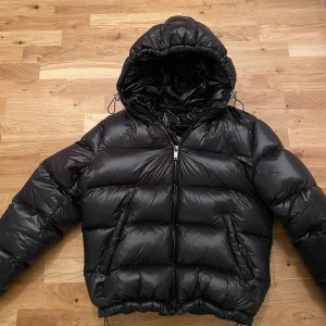 Svart pufferjacka Zara - Säljer en svart pufferjacka. Jackan har en glansig yta och är perfekt för kalla dagar. Den har långa ärmar och justerbara dragskor vid huvan för extra skydd mot väder och vind.                                                Jackan är köpt från Zara förra året men bara blivit använd 1 månad. ❗️Den är riven vid den högra fickan (bild 2) men annars är den i bra skick❗️