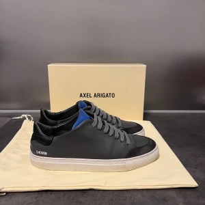 Axel Arigatos - Snygga grå sneakers från Axel Arigato med grå snörning och en vit sula. Skorna har en diskret blå detalj. Perfekta för en stilren look. Skorna är använda men i väldigt fint skick. Nypris ca 2500kr😀