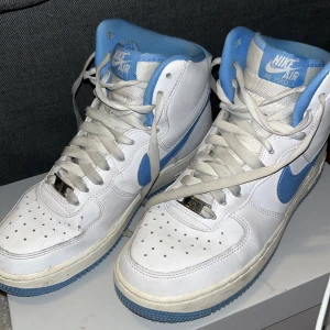 Nike Air Force 1 High i vitt och blått - Säljer ett par klassiska Nike Air Force 1 High sneakers i vitt med blå detaljer. Skorna har snörning och en hög design som ger extra stöd. Perfekta för en sportig look. Dem är fåtal använda men inte tvättad därför dem ser skitig ut. Mycket bra kvalitet och sko kartongen följer med.