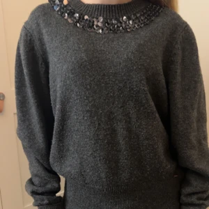 Mörkgrå tröja med paljettdetaljer - Snygg mörkgrå tröja med glittriga paljetter runt halsringningen. Tröjan har långa ärmar och ribbade muddar vid ärmslut och nederkant. Perfekt för att ge lite extra stil till din outfit.