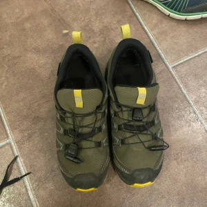 Gröna Salomon sneakers med gula detaljer - Säljer ett par gröna Salomon sneakers i mycket bra skick. De har gula detaljer och en robust sula perfekt för utomhusaktiviteter. Skorna har praktisk snörning och är tillverkade i slitstarkt material. Perfekta för äventyr i naturen!