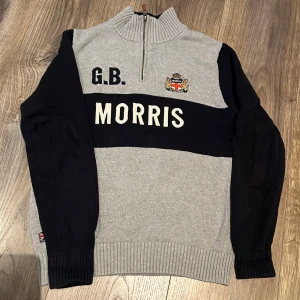 Grå och marinblå tröja från Morris - Säljer en snygg grå och marinblå tröja från Morris med dragkedja vid halsen. Den har ett broderat emblem och texten 'MORRIS' på bröstet. Perfekt för höst och vinter med sin stickade design och långa ärmar. Tröjan har ribbade muddar och är i mycket bra skick.