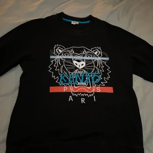 Svart sweatshirt från Kenzo - Säljer en svart sweatshirt från Kenzo med ett coolt tigertryck och texten 'Kenzo Paris' i blått och rött. Tröjan är i bra skick och perfekt för både vardag och fest. Den har långa ärmar och en normal passform. Passar perfekt för en avslappnad men uppklädd look.