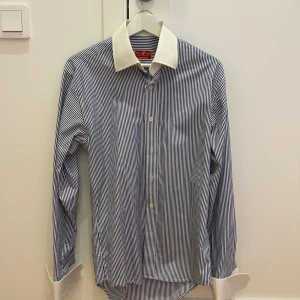 Randig skjorta från Tailor Store - Säljer en elegant randig skjorta från Tailor Store i blått och vitt. Skjortan har långa ärmar och vita kontrastkragar och manschetter. Perfekt för både formella och semi-formella tillfällen. Materialet känns som bomull och ger en skön passform. Passar bra till både kontoret och middagar. 🕴️