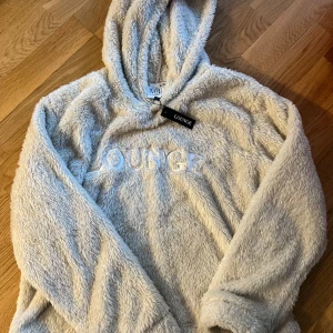 Beige fluffig hoodie från Lounge - Mysig och supermjuk beige hoodie från Lounge i storlek M. Perfekt för kalla dagar när du bara vill mysa hemma. Den har en stor luva och är gjord i ett fluffigt material som känns som en kram. Helt ny med tag kvar! 🧸