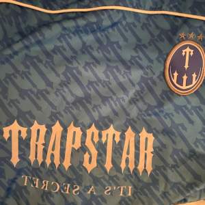 Säljer en blå fotbollströja från Trapstar med ett coolt kamouflagemönster. Var lite stor för migTröjan har korta ärmar och ett stort tryck med siffran 22 på baksidan. Märkets logga och texten 'It's a Secret' finns på framsidan. Perfekt för dig som vill sticka ut på planen eller gatan! 💙