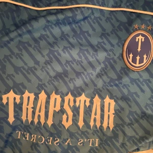  Trapstar Jersey  - Säljer en blå fotbollströja från Trapstar med ett coolt kamouflagemönster. Var lite stor för migTröjan har korta ärmar och ett stort tryck med siffran 22 på baksidan. Märkets logga och texten 'It's a Secret' finns på framsidan. Perfekt för dig som vill sticka ut på planen eller gatan! 💙