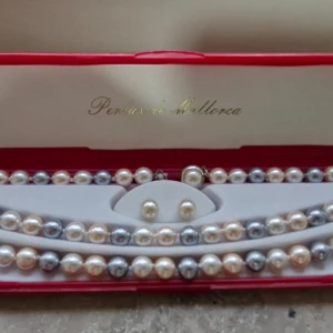 Pärlset från Perlas de Mallorca - Elegant pärlset från Perlas de Mallorca med halsband, armband och örhängen. Pärlorna är i vackra nyanser av vitt, silver och ljusrosa, vilket ger en klassisk och tidlös look. Perfekt för speciella tillfällen eller som en lyxig present. Setet kommer i en fin röd ask.