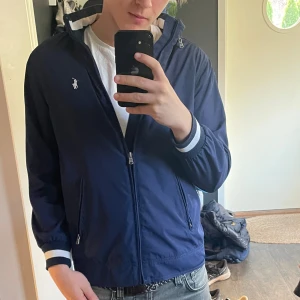 Mörkblå vindjacka från Ralph Lauren - Säljer en snygg mörkblå vindjacka från Ralph Lauren. Jackan har dragkedja och långa ärmar med vita detaljer vid ärmsluten. Perfekt för vår och höst! 🌀storlek L ungdom passar xs