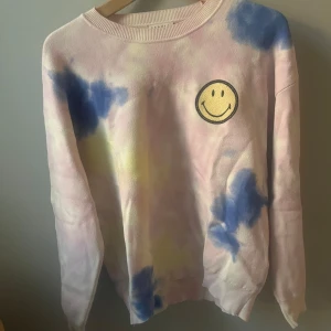 Tie-dye tröja med smiley - Supercool tie-dye tröja i pastellfärger med en glad smiley på bröstet. Perfekt för att liva upp garderoben! Tröjan har långa ärmar och en avslappnad passform. Perfekt för både vår och sommar.