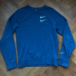 Sweatshirt Nike - Säljer en snygg blå sweatshirt från Nike med dubbla swoosh-loggor på framsidan och en stor text på baksidan. Tröjan har långa ärmar och är perfekt för både träning och vardag. Den är i mycket bra skick och superbekväm att ha på sig. Perfekt för höst och vår! Inga fläckar eller hål!