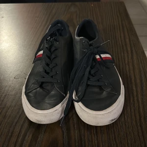 Svarta sneakers från Tommy Hilfiger - Säljer ett par svarta sneakers från Tommy Hilfiger i bra skick. De har en vit sula och snygga ränder i rött och vitt på sidan. Perfekta för vardagsbruk och har en cool stadssilhuett på sulan. Passar till både jeans och shorts! Kommer ej med box