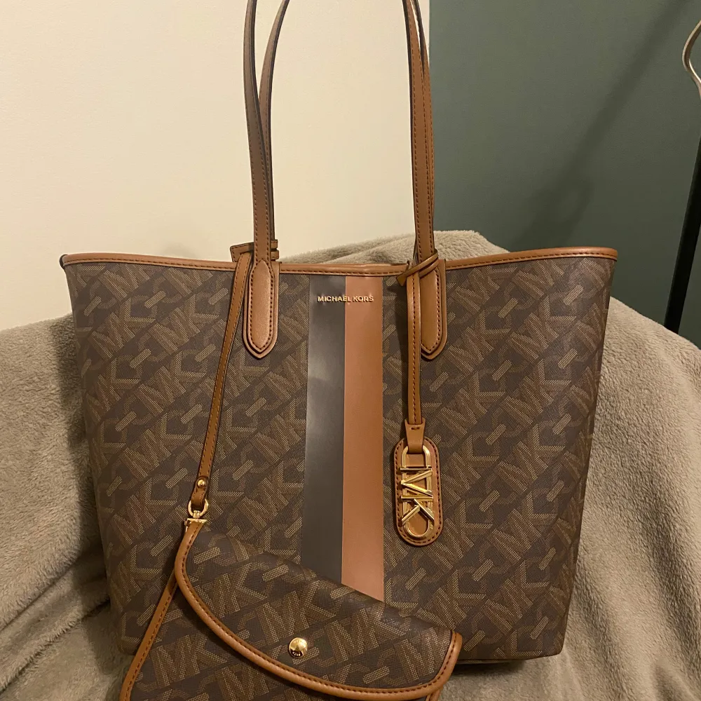 Säljer en äkta snygg brun Michael Kors Eliza Totebag i skinn med matchande plånbok. Väskan har ett stilrent mönster och detaljer i guld. Perfekt för både vardag och fest! Jag köpte den i år och har använt bara 2 gånger.. Väskor.