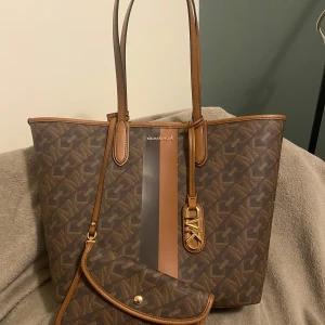 Brun Michael Kors Eliza Totebag i skinn - Säljer en äkta snygg brun Michael Kors Eliza Totebag i skinn med matchande plånbok. Väskan har ett stilrent mönster och detaljer i guld. Perfekt för både vardag och fest! Jag köpte den i år och har använt bara 2 gånger.