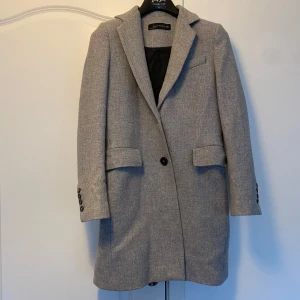 Grå kappa från Zara Woman - Säljer en stilren grå kappa från Zara Woman i storlek S. Den har en klassisk design med långa ärmar och knappar på ärmsluten. Perfekt för höst och vår, och ger en elegant touch till vilken outfit som helst. Kappan är i mycket bra skick och redo för nya äventyr!