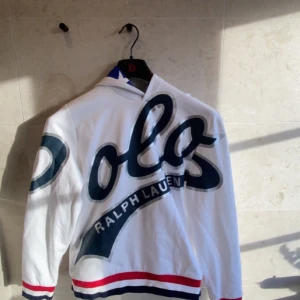 Vit hoodie från Ralph Lauren - Säljer en snygg vit hoodie från Ralph Lauren med stor blå logga på framsidan. Den har en cool stjärnmönstrad insida i luvan och röda samt blå ränder vid ärmslut och nederkant. Perfekt för en avslappnad stil!