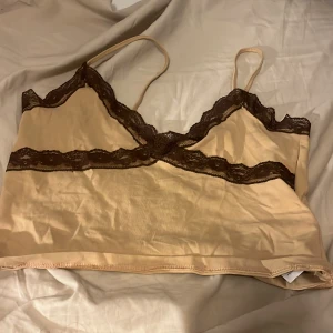 Beige linne med spetsdetaljer från New Yorker - Säljer ett snyggt beige linne från New yorker i storlek M. Det har tunna axelband och fina bruna spetsdetaljer längs kanterna. Perfekt för sommardagar eller som ett lager under en tröja.