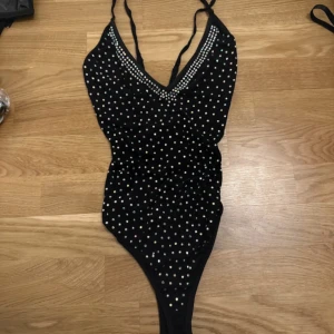 Svart body med prickigt mönster - Snygg svart body med vita prickar och smala axelband. Perfekt för fest eller poolhäng. Den har en v-ringad design som ger en stilren look. body är i mycket bra skick och passar perfekt för fest eller vardagsliv 