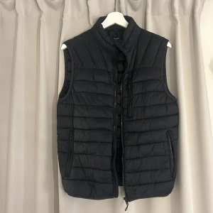 Svart dunväst från Bershka - Säljer en svart dunväst från Bershka i storlek S. Västen har en dragkedja framtill och två praktiska fickor med dragkedjor. Perfekt för höst och vår när du vill hålla dig varm men ändå se stilren ut. Passar bra till både vardag och lite mer avslappnade tillfällen.