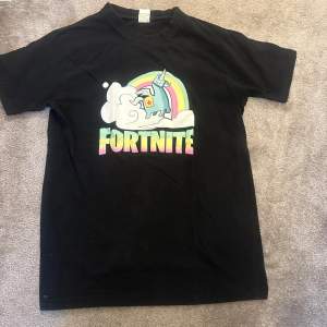 Säljer en svart Fortnite t-shirt i 100% mjuk bomull. Den har ett färgglatt tryck med en enhörning och regnbåge på framsidan. Perfekt för gaming eller casual wear. Skön och bekväm passform!