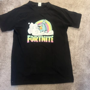Svart Fortnite t-shirt - Säljer en svart Fortnite t-shirt i 100% mjuk bomull. Den har ett färgglatt tryck med en enhörning och regnbåge på framsidan. Perfekt för gaming eller casual wear. Skön och bekväm passform!