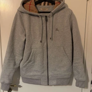Grå hoodie från Burberry - Säljer en stilren grå hoodie från Burberry i storlek S. Den har en klassisk dragkedja och en mysig huva med det ikoniska Burberry-mönstret på insidan. Perfekt för både vardag och avslappnade tillfällen. Huvan har dragsko och tröjan har praktiska fickor framtill.