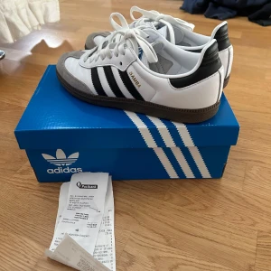 Adidas Samba sneakers - Säljer ett par klassiska Adidas Samba sneakers i vitt och svart. Skorna har en snygg design med svarta ränder och grå detaljer i mocka. De är i nyskick och kommer med originalkartong. Perfekta för både vardag och träning!
