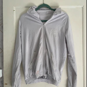 Vit jacka med broderad krona - Säljer en stilren vit jacka med dragkedja och broderad krona på bröstet och ryggen. Jackan har långa ärmar och ribbade muddar vid ärmslut och nederkant. Perfekt för vår och höst!