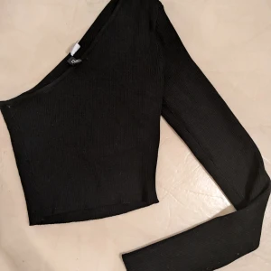 Svart oneshoulder topp - Säljer en snygg svart oneshoulder topp som är perfekt för festliga tillfällen. Den är ribbad och har en lång ärm som ger en cool asymmetrisk look. Toppen är tight men med mycket stretch. Bara använd 1 gång.