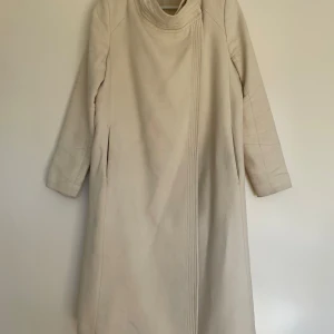 Beige kappa från Whyred - Säljer en stilren varmvit/ ivoryvit kappa från Whyred. Den har en elegant design med en hög krage och knäpps med en knapp. Bullstyg och fodrad - Perfekt för höst och vår. Kappan är i bra skick men har spår av användning i form av svagare fläckar, dock ej påtagliga när den bärs. Har därav satt ett lågt pris! 