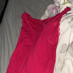 Röd ribbad linne med spetsdetaljer - Säljer ett snyggt mörkrosa ribbat linne med spetsdetaljer vid halsringningen och ärmarna. Perfekt för sommardagar eller som ett lager under en kofta. Linnet är ärmlöst och stretchigt.