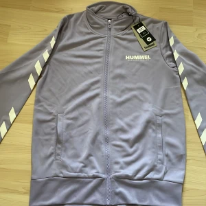 Lila träningsjacka från Hummel - Säljer en snygg lila träningsjacka från Hummel (ljusare pastel i verkligen)💜.Jackan har en dragkedja framtill och vita detaljer på ärmarna. Den är långärmad och har två fickor på framsidan. Perfekt för träning eller vardagsbruk. Helt ny med taggar kvar!