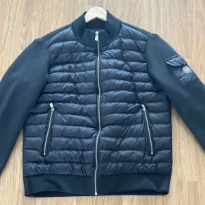 Svart quiltad jacka från Moncler - Säljer en snygg svart quiltad jacka från Moncler. Den har en stilren design med dragkedja framtill och två praktiska fickor med dragkedjor. Jackan har långa ärmar och en cool patch på ena ärmen. Perfekt för höst och vår!
