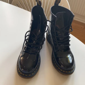 Svarta lackade kängor från Dr. Martens - Säljer ett par svarta, glansiga kängor från Dr. Martens i storlek 38. De har den klassiska gula sömmen och snörning framtill. Perfekta för höst och vinter med sin robusta design och slitstarka material. Använda men i bra skick!