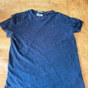 Blå t-shirt från Tommy Jeans - Säljer en snygg blå t-shirt från Tommy Jeans i storlek M. Den har en regular fit och är kortärmad, perfekt för vardagsbruk. T-shirten är i bomull och har en liten broderad logga på bröstet. Passar bra till både jeans och shorts!🔥