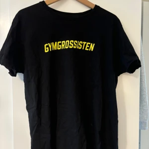 Svart t-shirt från Gymgrossisten - Säljer en svart t-shirt från Gymgrossisten i storlek XL. Den har en enkel design med texten 'GYMGROSSISTEN' i gult på framsidan. Perfekt för träning eller vardagsbruk! T-shirten är i nyskick!