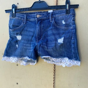 Blå jeansshorts med spetsdetalj - Säljer ett par snygga blå jeansshorts från &Denim. De har en cool distressed look med slitningar och en söt spetskant längst ner. Perfekta för sommaren och passar både till stranden och en dag på stan. De är i bra skick och redo för en ny ägare! 