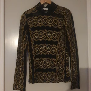 Svart och guldmönstrad topp från New Look - Säljer en elegant svart topp från New Look med ett snyggt guldmönster. Toppen är långärmad och har en tight passform, perfekt för festliga tillfällen.  Liten i storleken