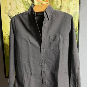 Grå skjorta från J.Lindeberg - Säljer en stilren grå skjorta från J.Lindeberg. Skjortan har en klassisk design med knappar framtill och en bröstficka. Perfekt för både vardag och festliga tillfällen. Den är i bra skick och passar perfekt till höst- och vårgarderoben.